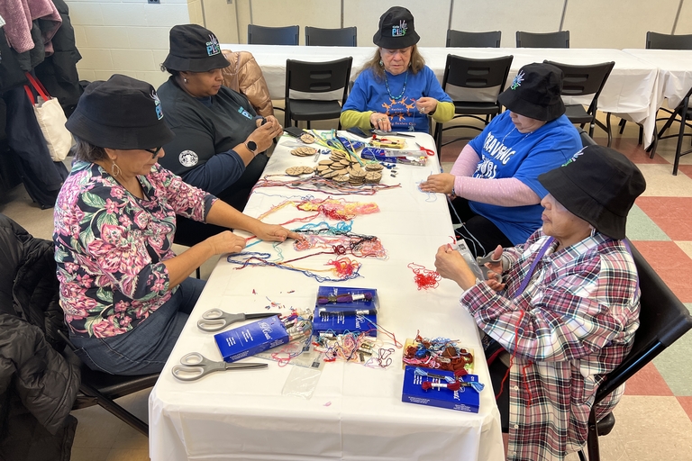Five women sit around a table working with colorful embroidery thread and cardboard circles.