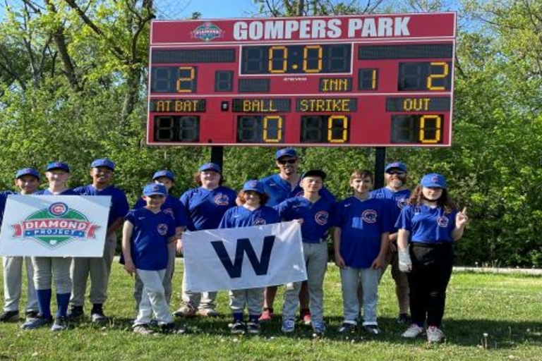 Youth baseball team and coaches pose with a "W" banner under the Gompers Park scoreboard.
