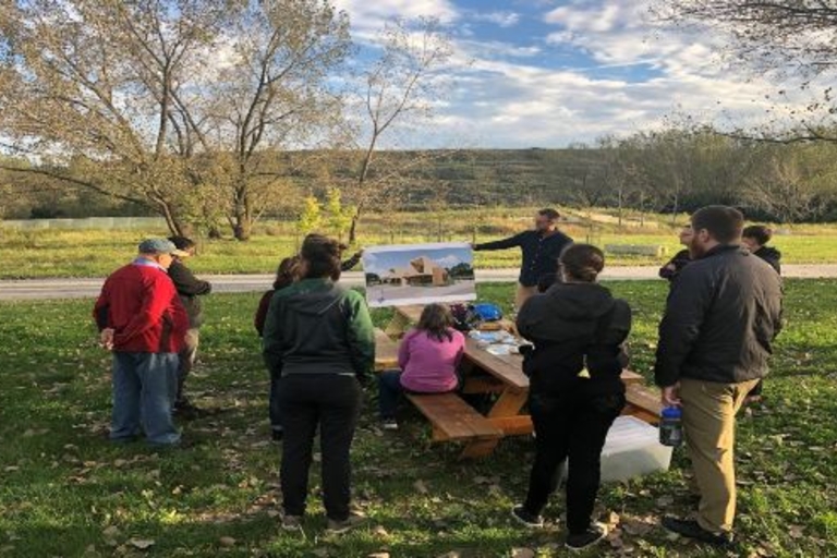 Group gathers outdoors around picnic table reviewing architectural drawing.