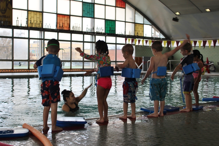 Children with flotation devices stand at edge of pool as instructor waits in water.
