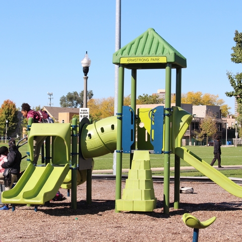 Green and blue playground structure at Armstrong Park. Children play on the equipment and others gather nearby on a sunny day.
