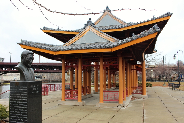 Chinese-style pavilion with tiered, tiled roof and red accents beside a bust and river.