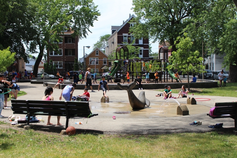 Children play around a whale sprinkler in a park on a sunny day.