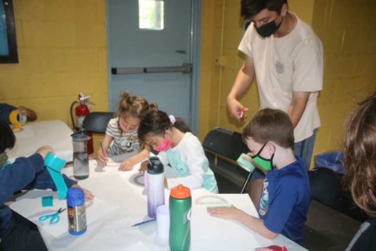 Children wearing masks do crafts at a table.  A masked adult supervises.