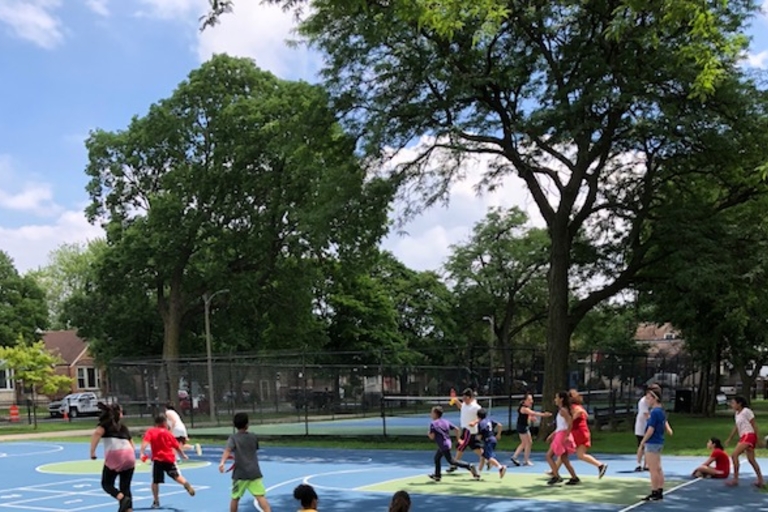Children playing on a blue and green outdoor basketball court on a sunny day. Surrounding trees and a partially cloudy sky.