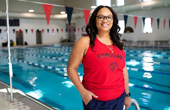A lifeguard in a red lifeguard tank top stands by an indoor pool.