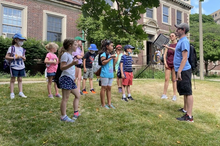 A group of children listens to two tour guides on a grassy area in front of a brick building.