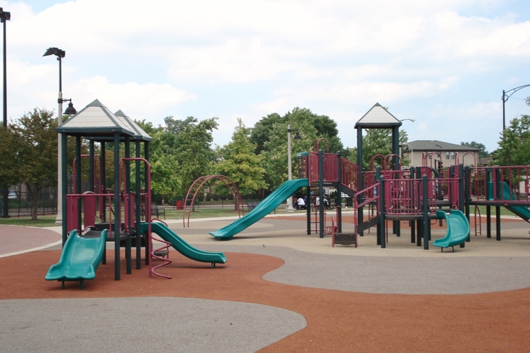 Playground with green and red structures and slides on a mottled surface of gray and rust-colored soft material.