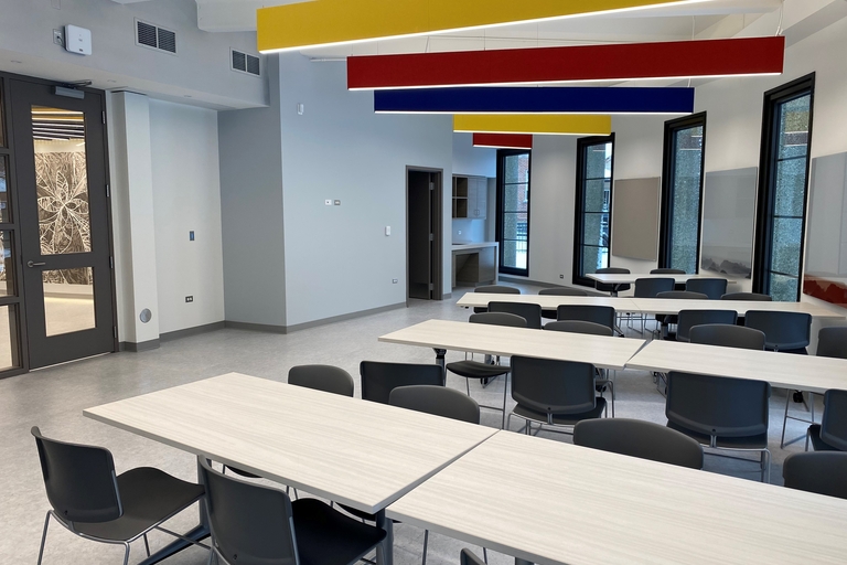 Modern classroom with light wood tables and dark gray rolling chairs. Colorful, rectangular lights hang from the ceiling.