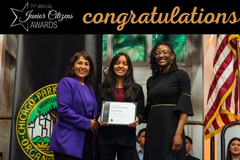 Three women at an awards ceremony, one holding an award. An American flag and Chicago Park District banner are visible.