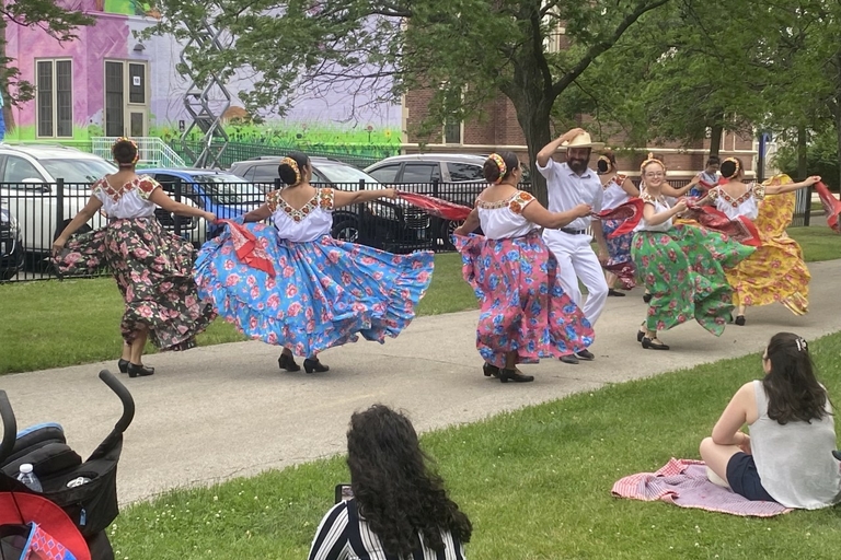 Folkloric dancers in colorful skirts and white tops perform outdoors. A person watches from the grass.

