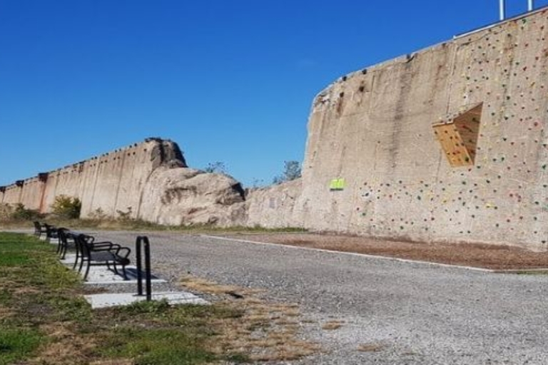 A concrete climbing wall with colorful handholds beside a long, low concrete wall. Benches sit on a paved area at the base of the wall.