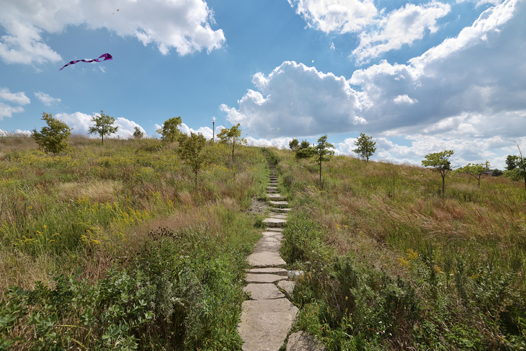 Stone path leads up grassy hill under cloudy, blue sky with kite flying.
