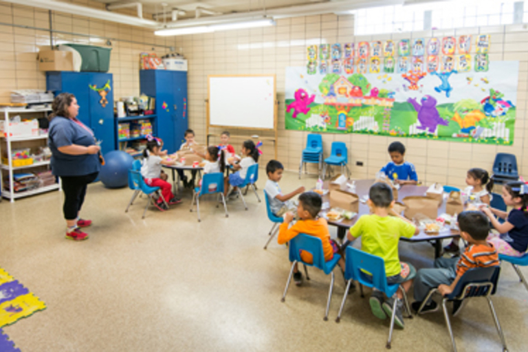 Preschool children eat lunch at tables while a teacher watches.