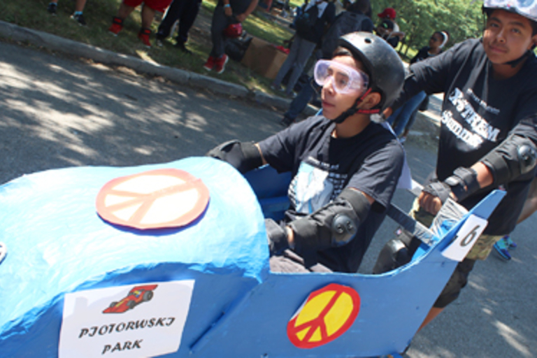 Child in blue go-cart being pushed by a teenager.