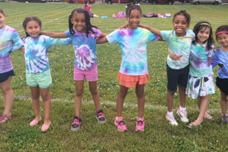 Eight children in tie-dye shirts stand arm in arm on a grassy field.
