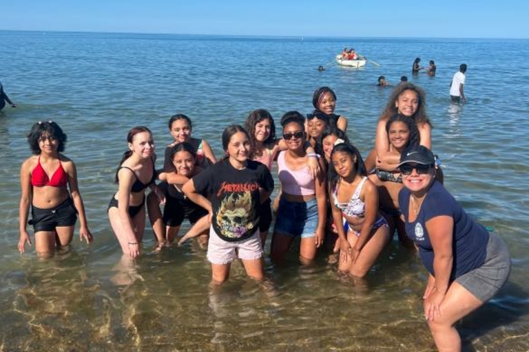 Group of girls and a chaperone pose in shallow ocean water on a sunny day.