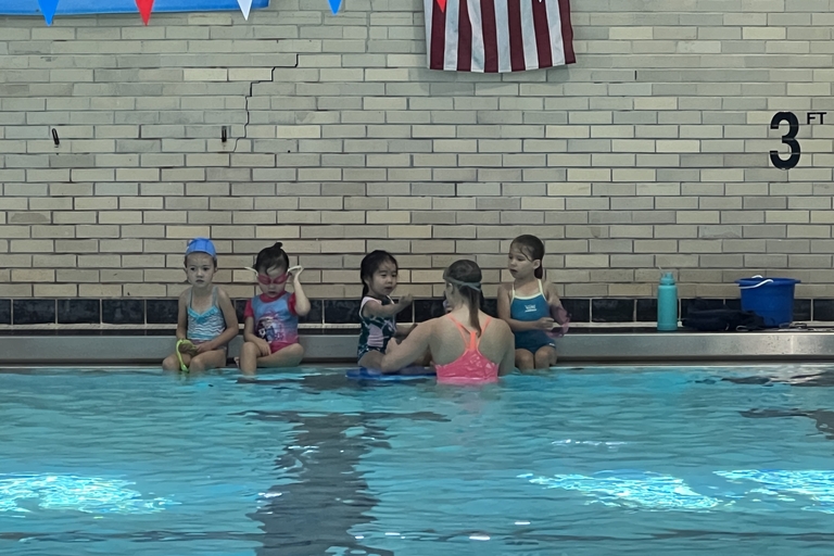 Four children and a swim instructor sit at the edge of a pool.