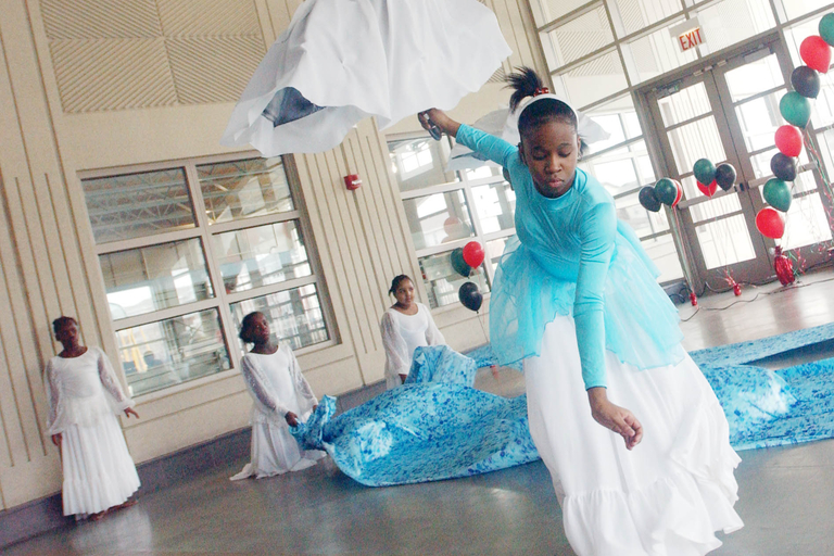A young girl in a teal and white dress dances with a white flowing prop. Other dancers in white dresses are visible.