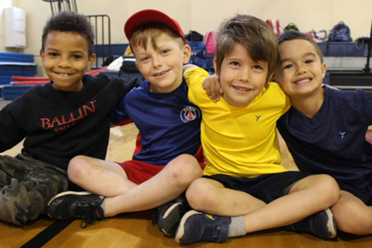 Four boys sit with legs crossed and arms around each other, smiling at the camera in a gym.