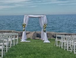 Lakefront wedding setup with white chairs and floral-decorated canopy.