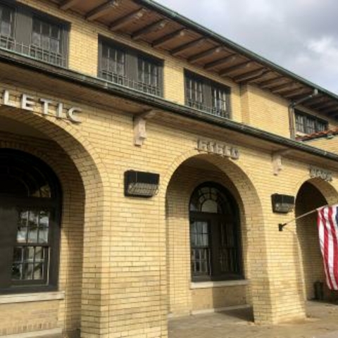 Exterior of a tan brick building with arched doorways and an American flag.  "Athletic" is written above one doorway.