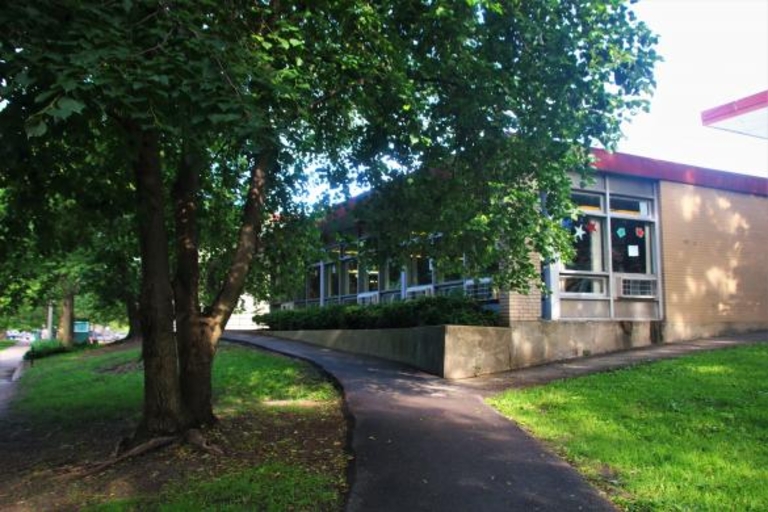 A low, long building with large windows sits behind a short retaining wall near a paved path and leafy trees.