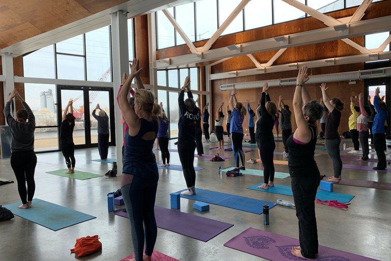 Large group performs yoga with arms raised in a studio with large windows and industrial views.
