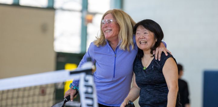 Two women smile at a pickleball net. One has her arm around the other.
