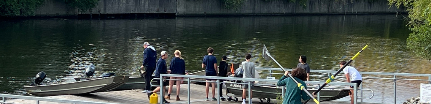 Rowing team and coaches gather at a dock with motorboats and oars.