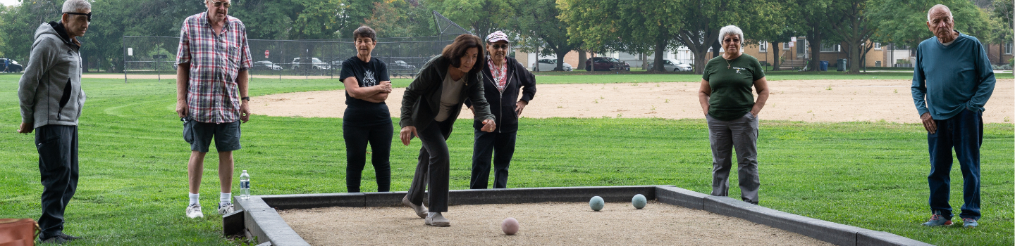 A group of people play bocce ball in a park. One player tosses a ball toward others already in the court.
