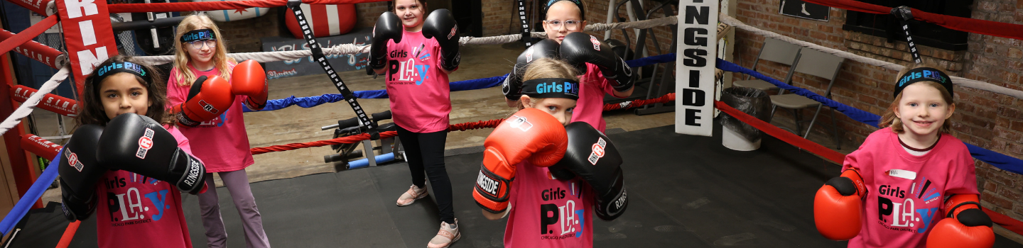 Five girls wearing boxing gloves stand in a boxing ring.