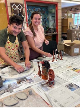 Two smiling people work on clay projects at a table covered in newspaper and small sculptures.