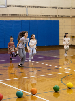 Four girls run on a gymnasium floor with dodgeballs lined up.