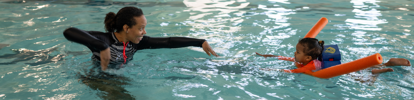 Adult teaches a toddler to swim in a pool. The child wears a flotation device and uses pool noodles.