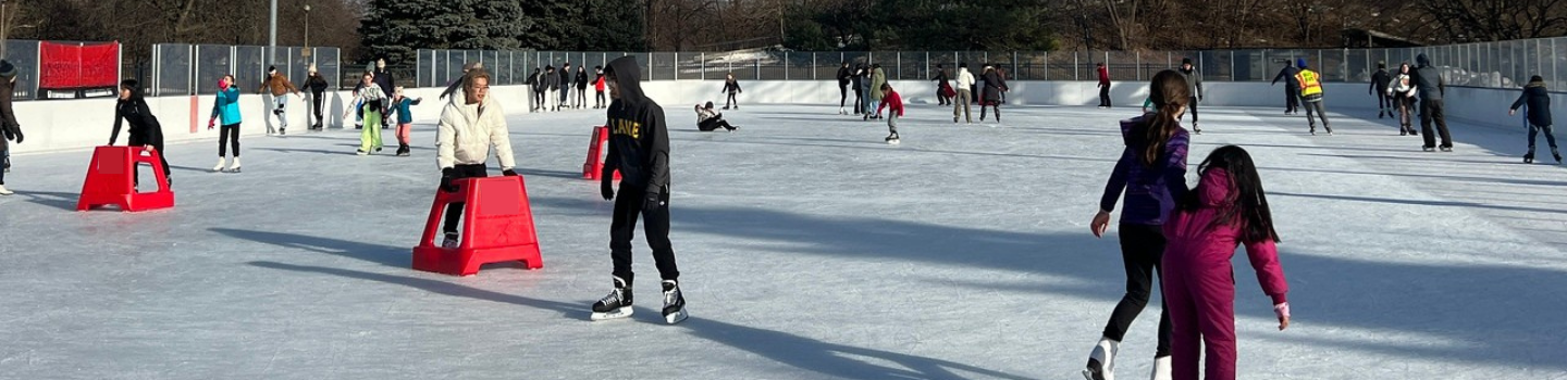 People ice skating at an outdoor rink. Some use red skating aids.