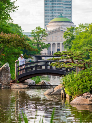 Woman on arched footbridge in tranquil Japanese garden. Building and skyscraper in background.