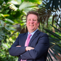 Smiling man in suit and tie, arms crossed, in front of foliage.