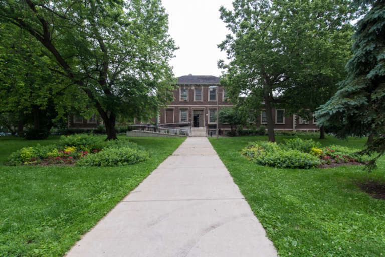 Photograph of brick building behind trees and sidewalk.