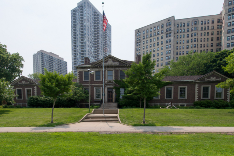 Brick building with U.S. flag, surrounded by park and highrises.
