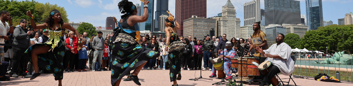 African dancers and drummers perform outdoors for a crowd, with a city skyline in the background.