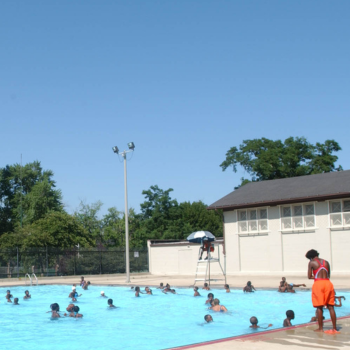 People swimming in a bright blue public pool on a sunny day. Lifeguard stands watch.