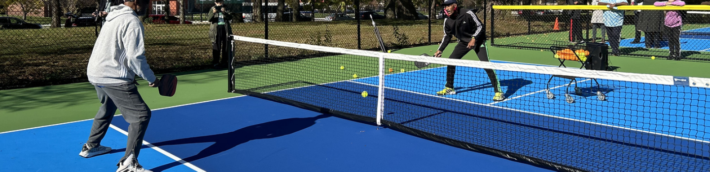 Two people play pickleball on a sunny day. Spectators watch from behind the fence.