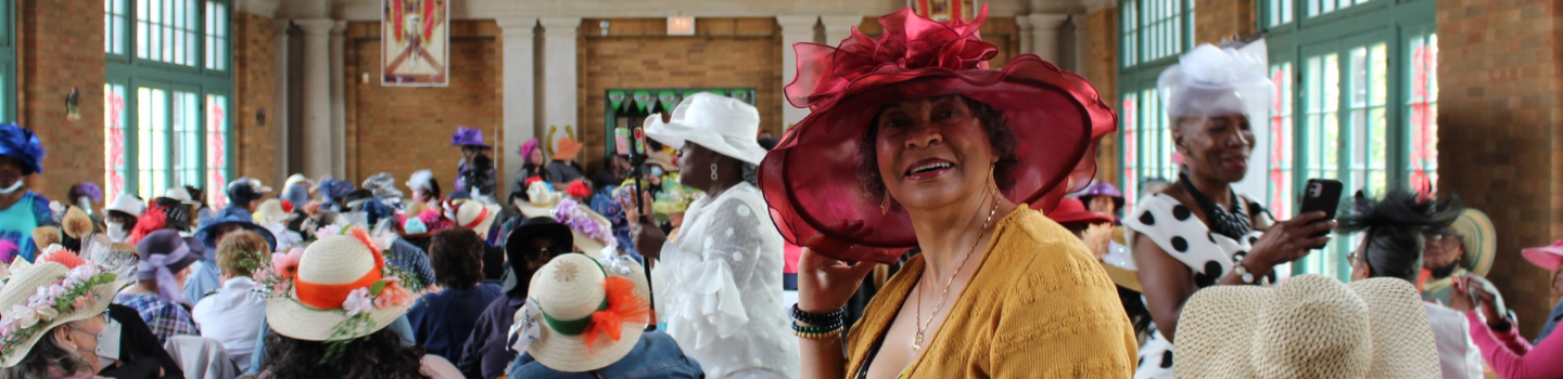 Woman in a maroon hat smiles at a gathering of women wearing decorated hats.