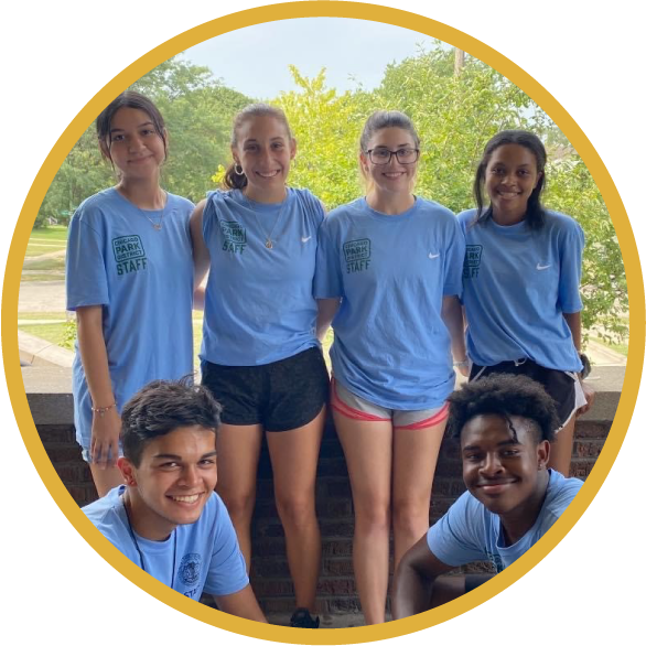 A group of six Recreation Leaders pose together for a photo, while in their park uniforms, outdoors in a park with trees in the background.
