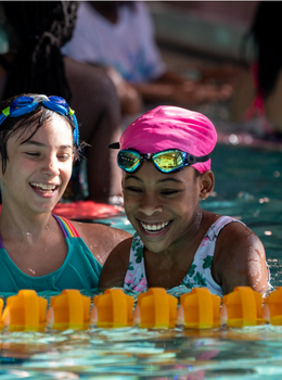Two smiling girls wearing swimsuits and goggles in a pool.